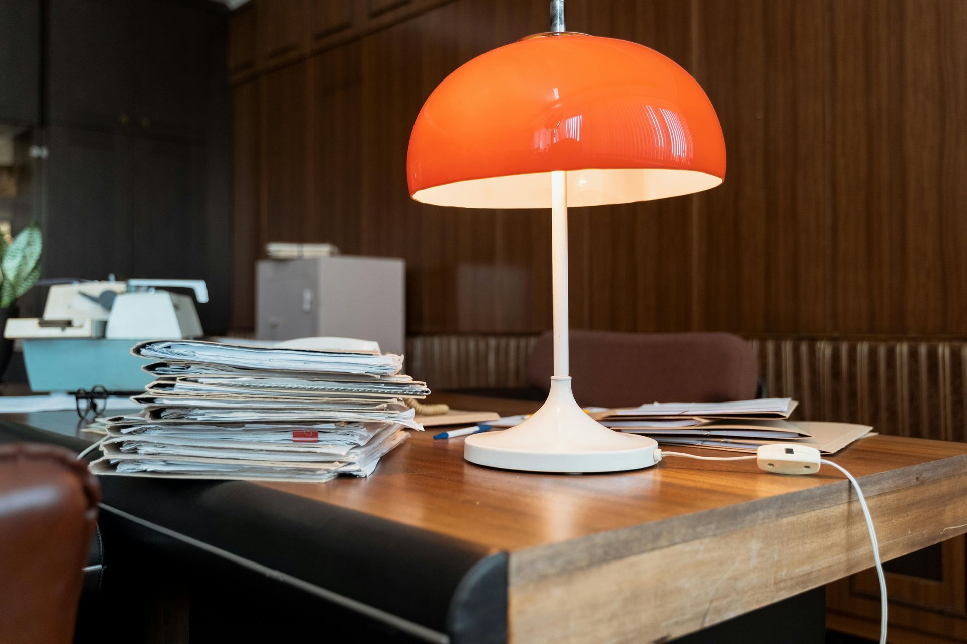 Een oranje lamp op een rommelig houten bureau met papieren en een typemachine op de achtergrond.
