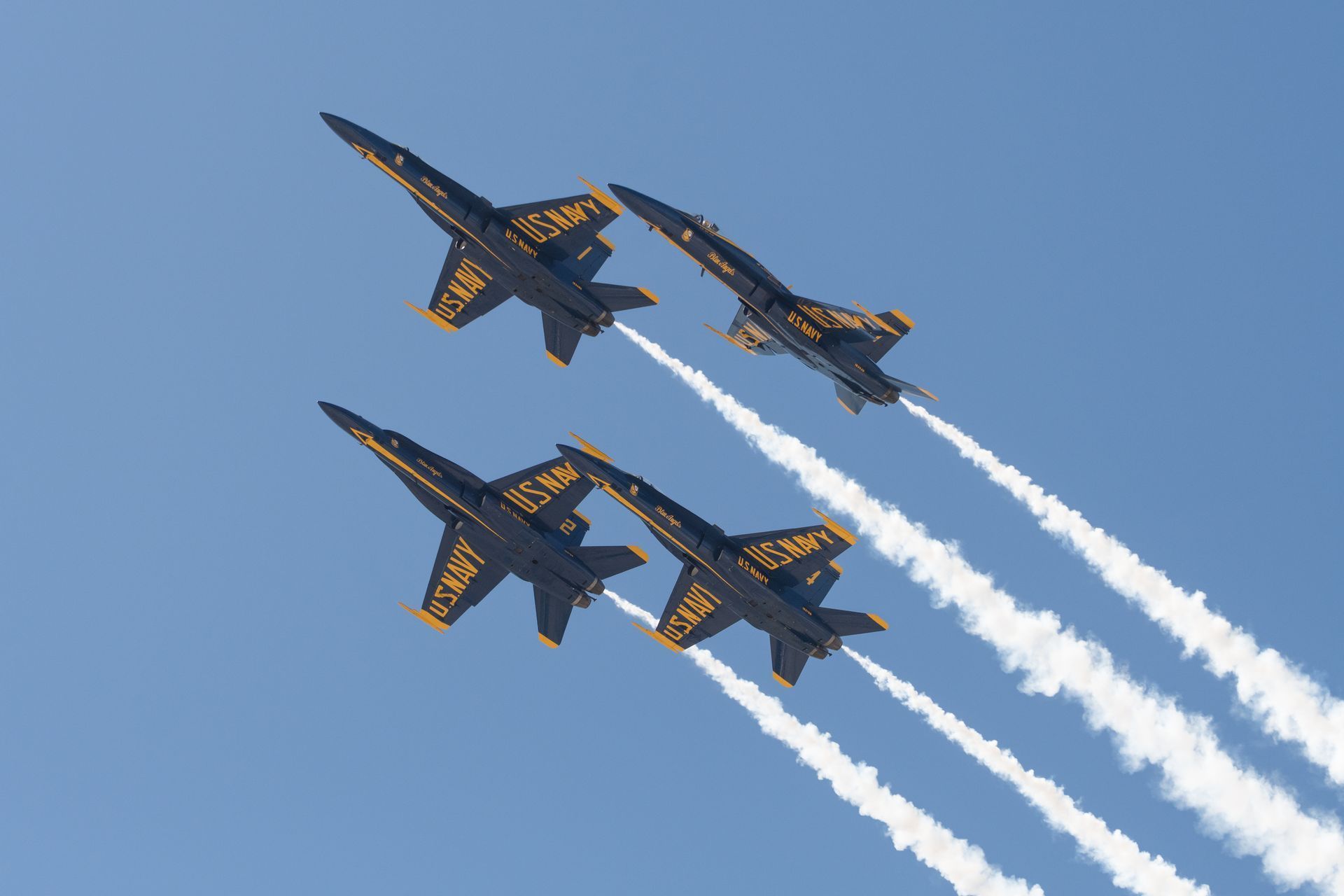 Four blue and yellow U.S. Navy Blue Angels jets flying in formation, leaving white contrails against a clear blue sky.