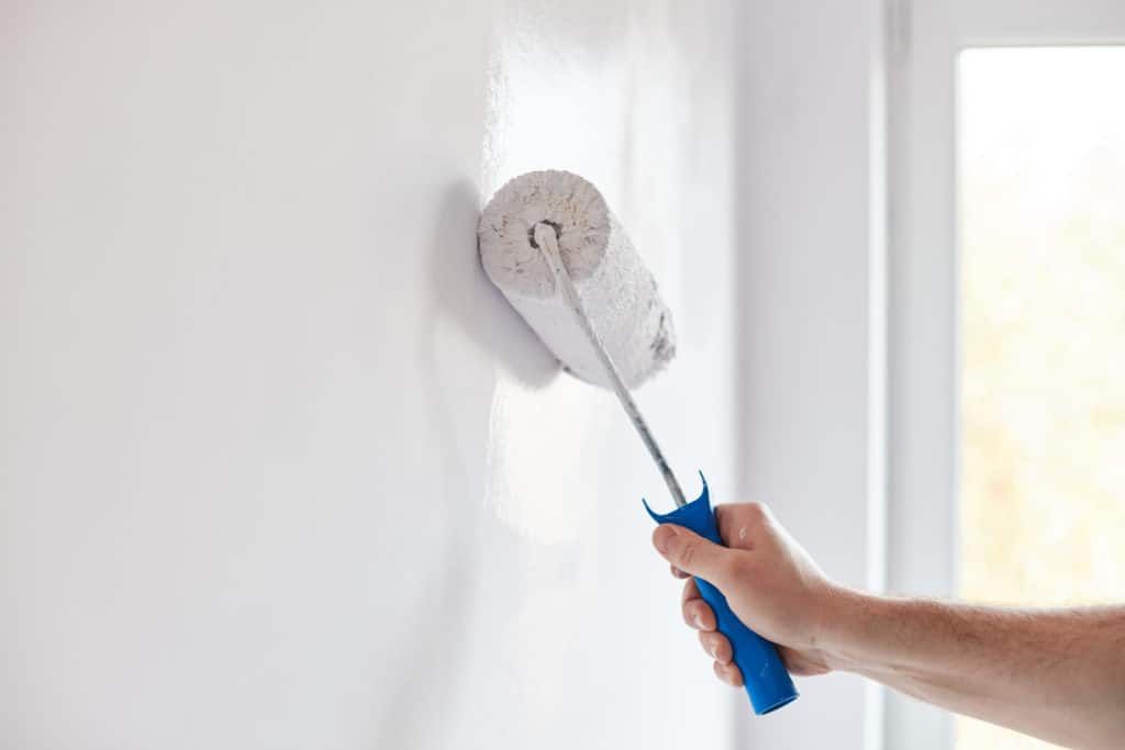 Hand painting a white wall with a paint roller in a room with a window.