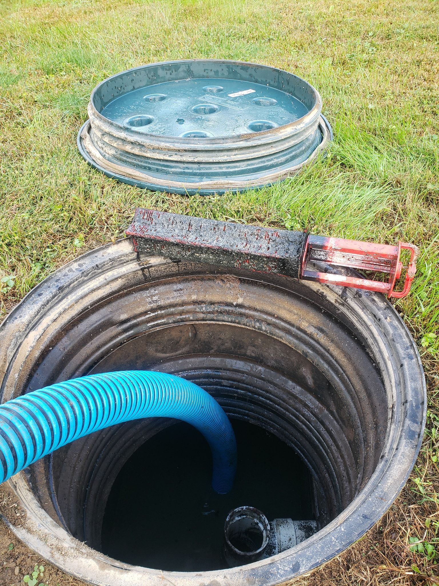 Septic tank being pumped with blue hose, lid off, in grassy yard.