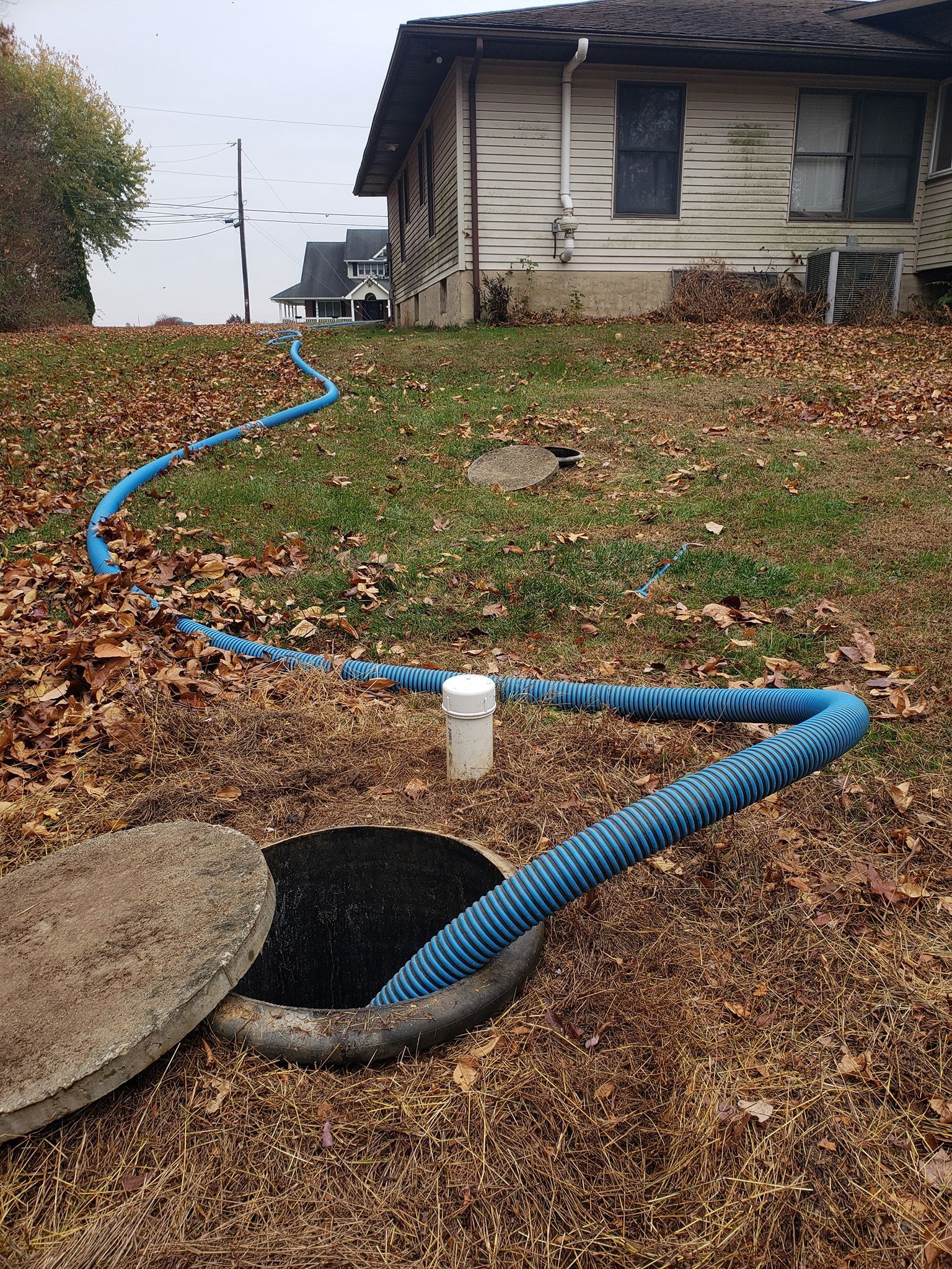 Open septic tank with blue hose, near a house covered in leaves, outdoor.