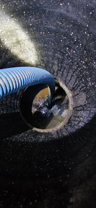 Worker with orange glove lowers pipe into an open sewer. Gray sewer grate and equipment visible.