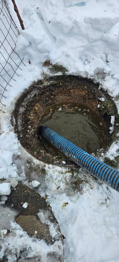Hose in open septic tank, removing waste. Green metal rim, brown hose, outdoor setting.