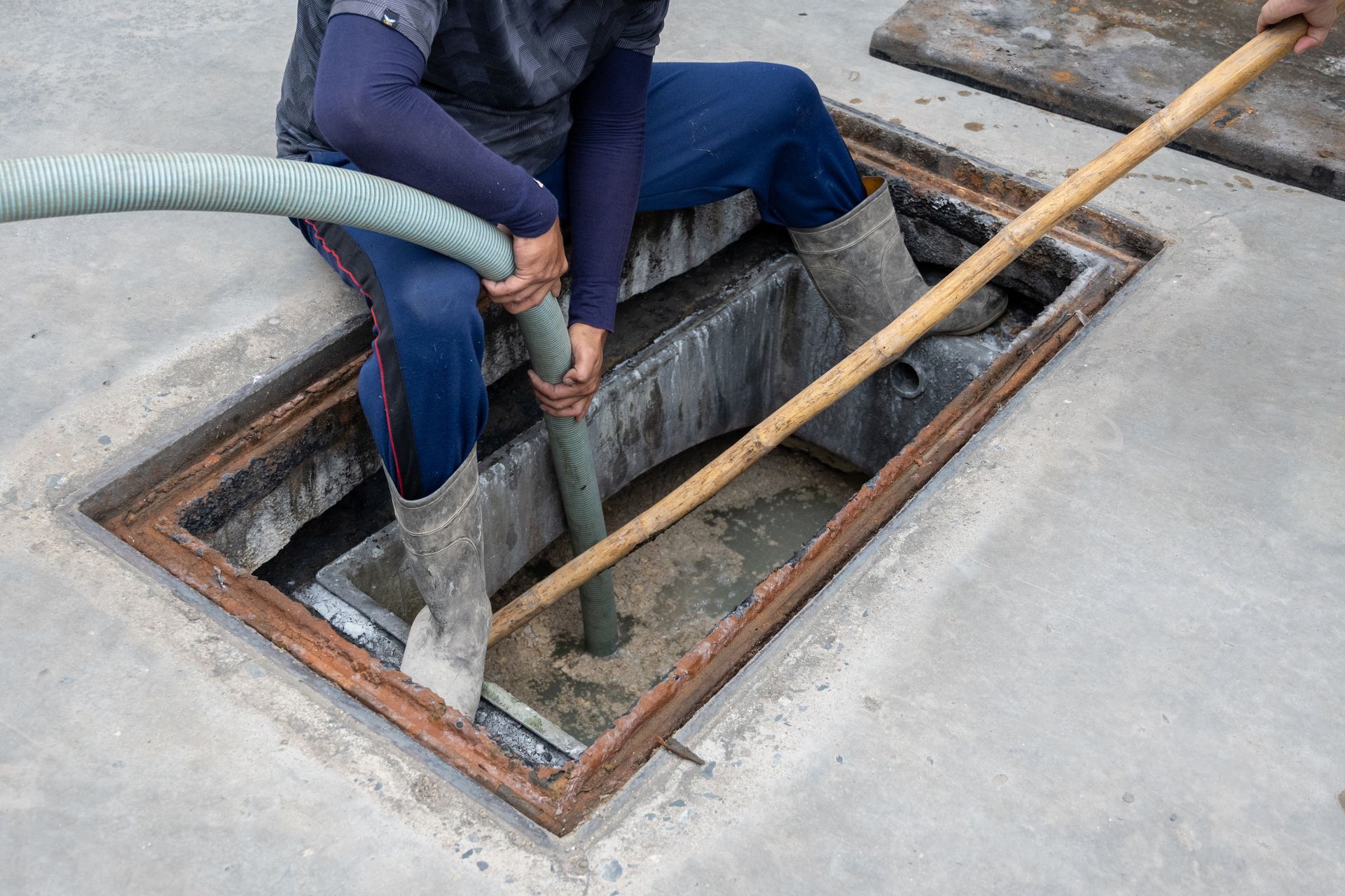 Person emptying a septic tank with a hose and shovel, seated at the opening. Person emptying a septic tank with a hose and shovel, seated at the opening.