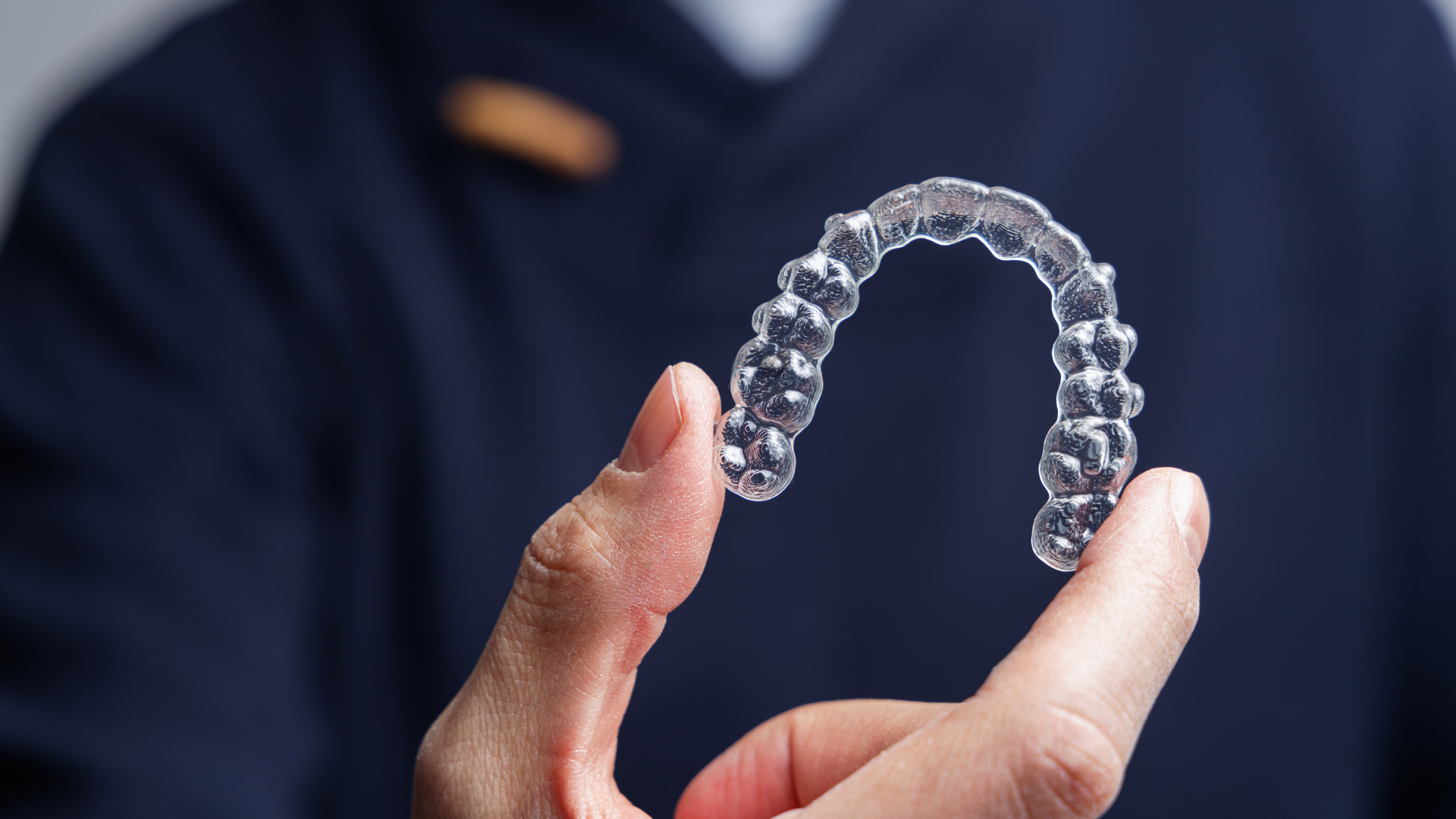 Clear orthodontic aligner held in hand against a dark blue background.
