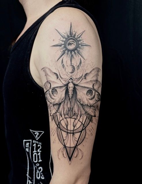 Tattoo of a moth with geometric shapes on arm, sun with eye above. Black ink on skin.