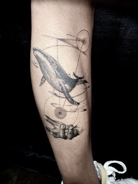 Tattoo of a whale, hand, and geometric shapes on an arm in black ink.