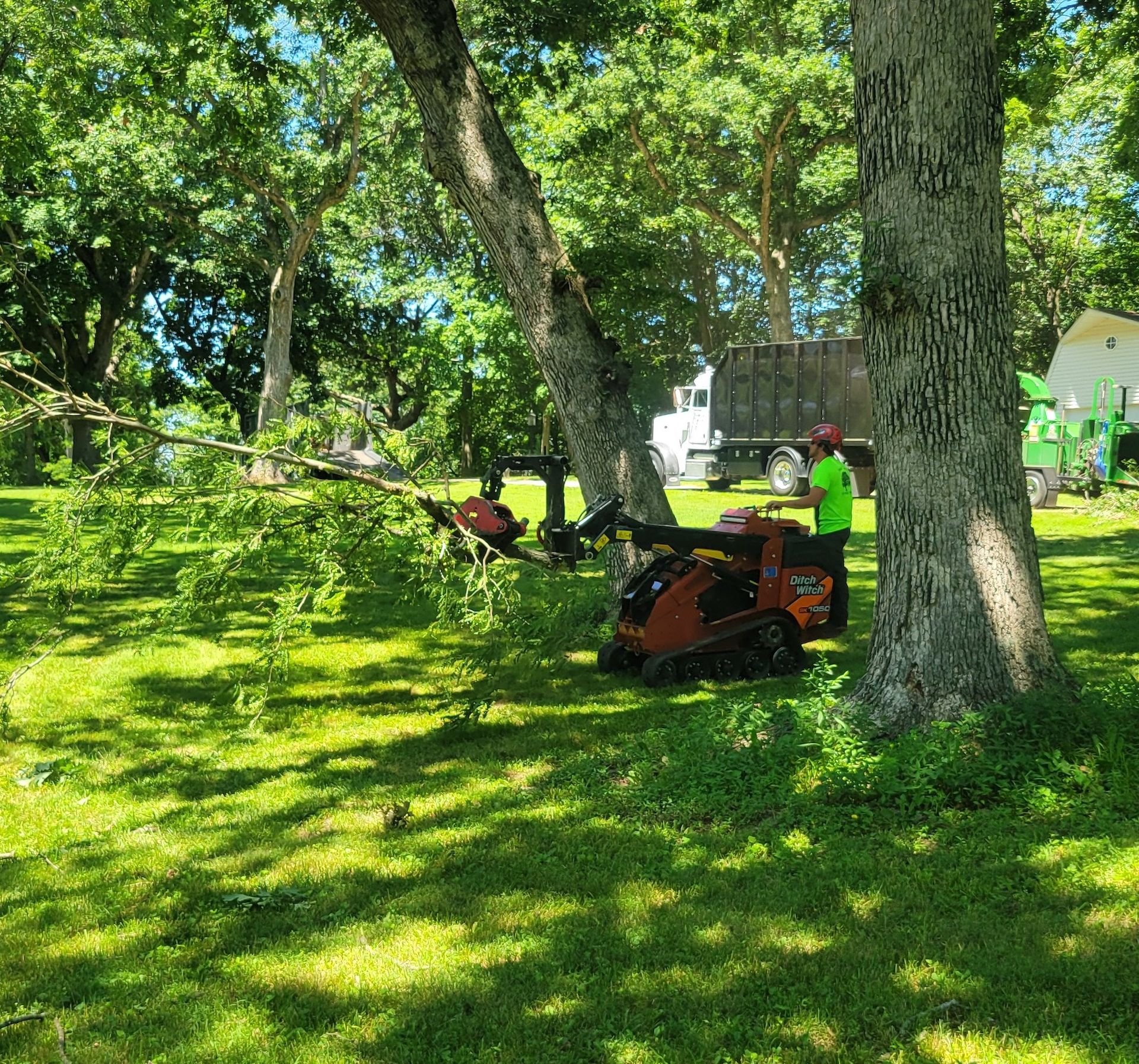 A worker in green shirt uses a small orange machine in a grassy yard, a truck and wood chipper in the background.