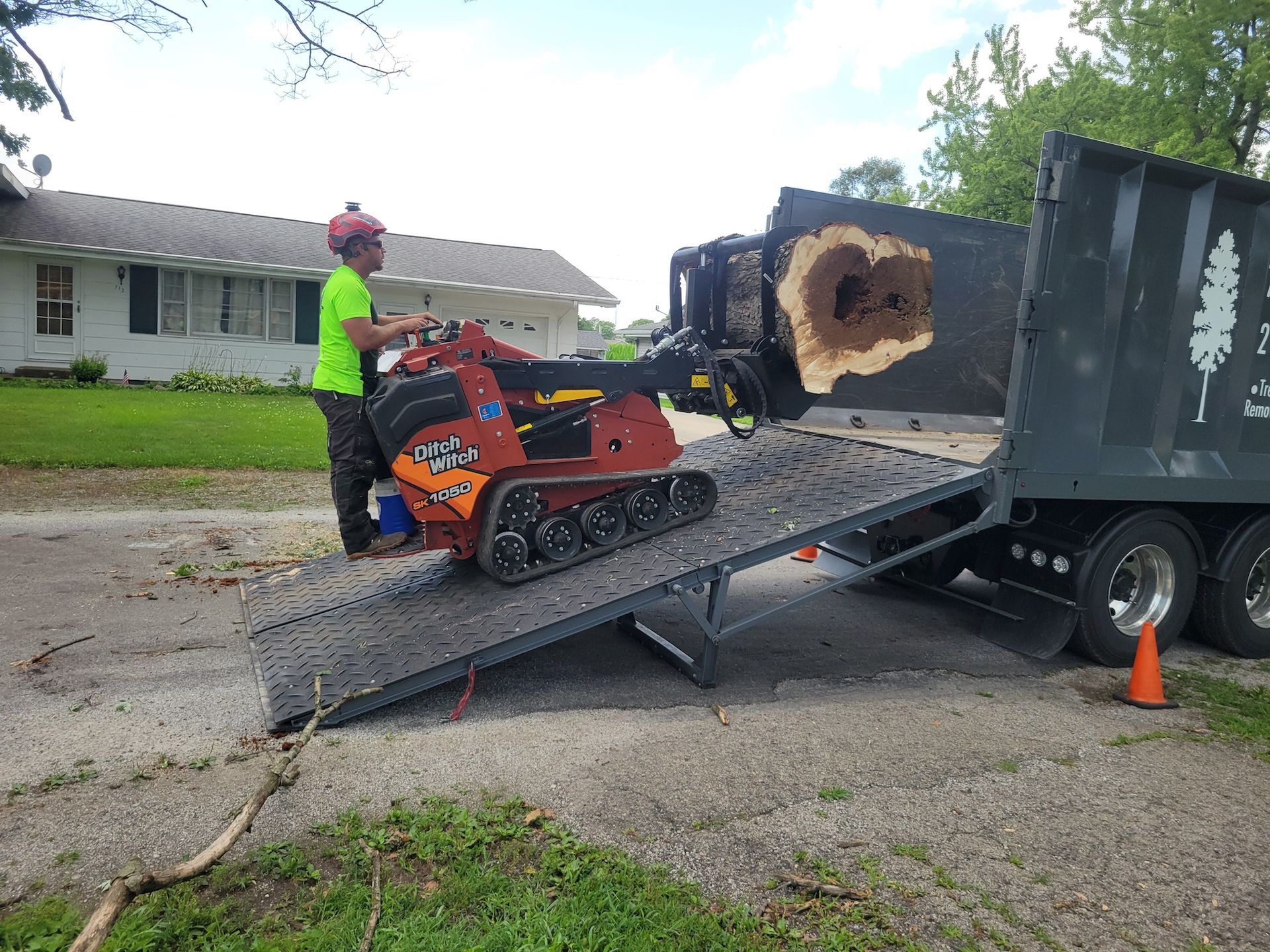 Man operating a mini skid steer loading wood chips into a truck, on a residential street.