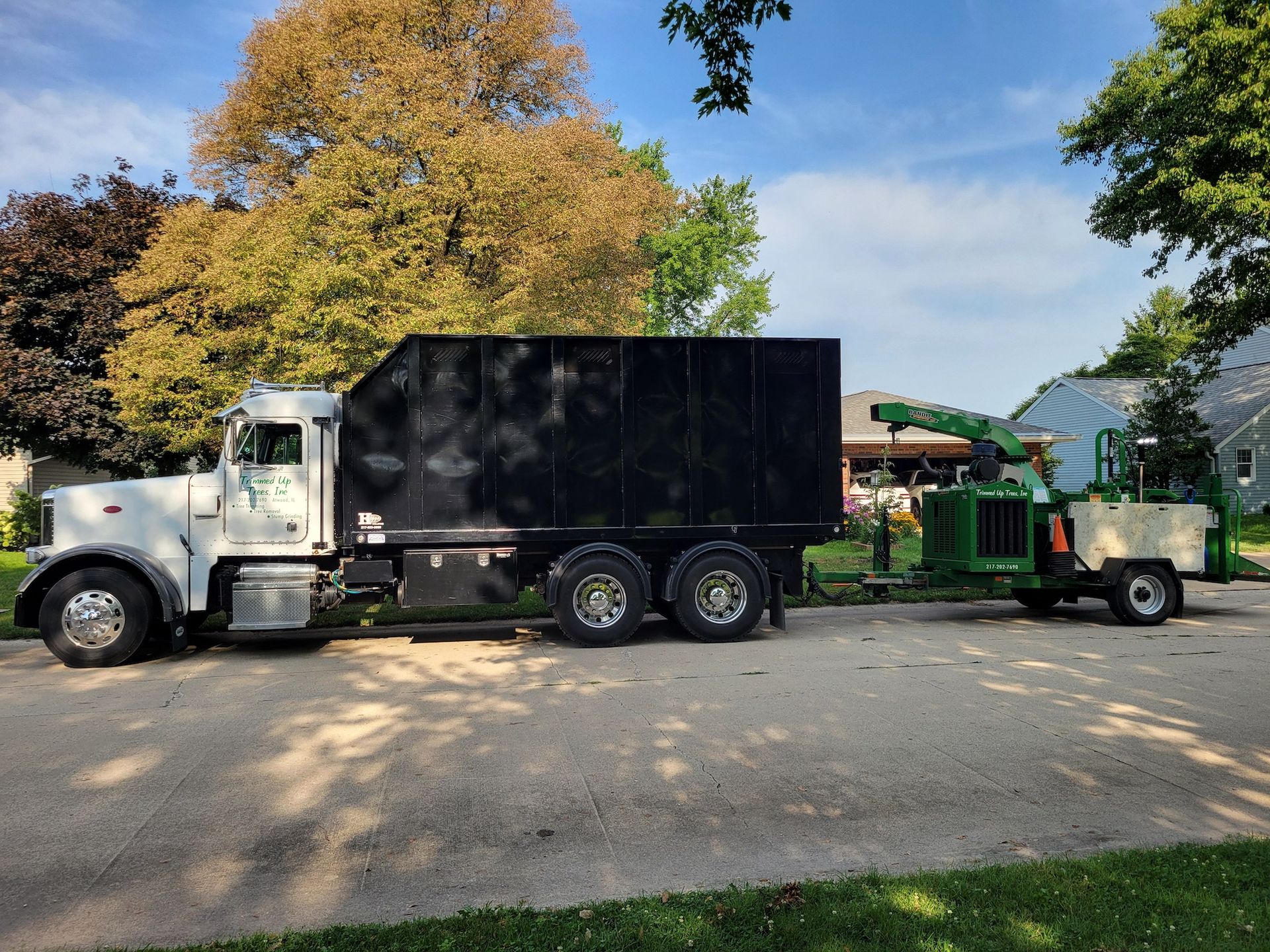 White truck with black container and a green wood chipper parked on a residential street.