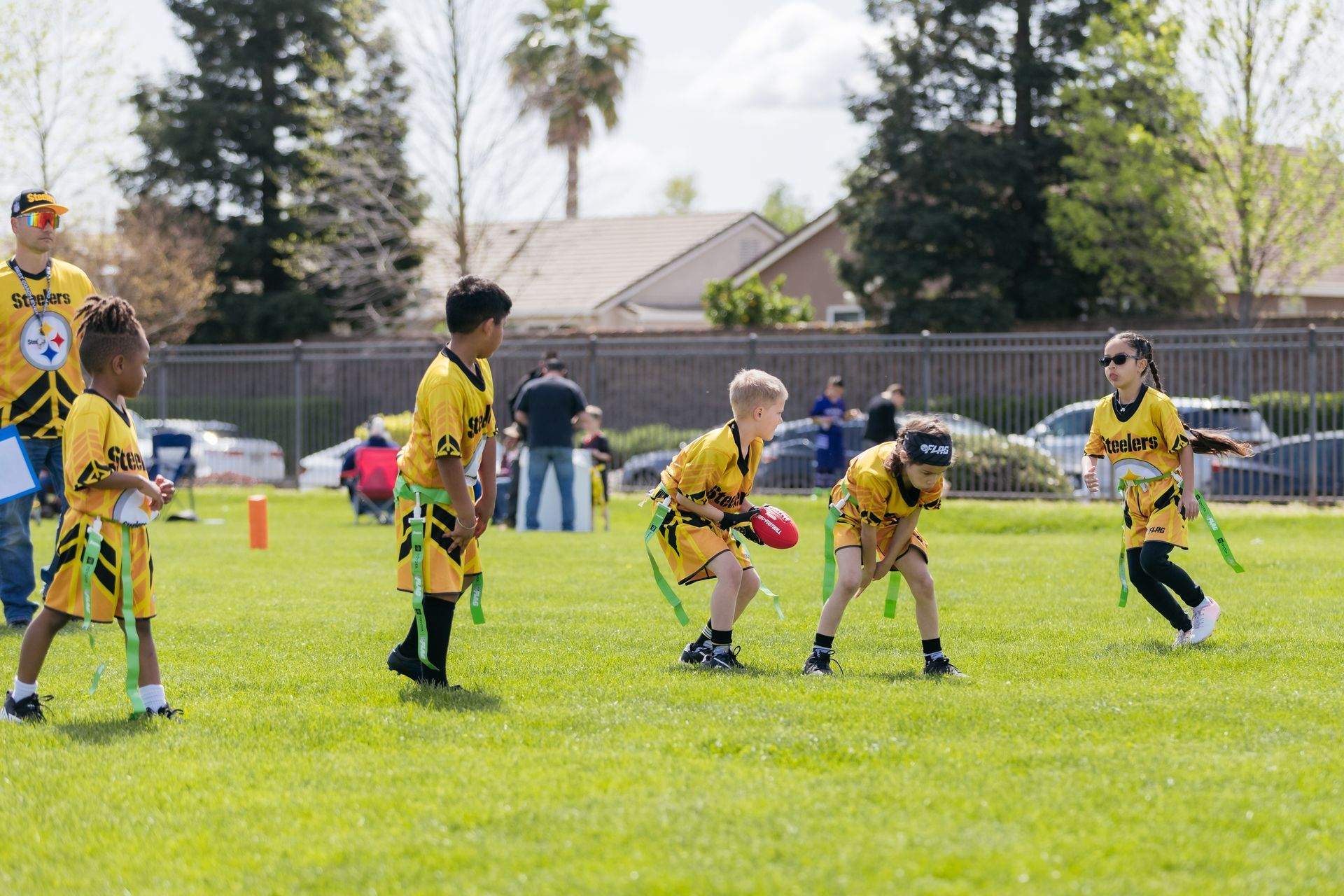 Children playing flag football on a grassy field, wearing yellow jerseys and flags, with a coach nearby.