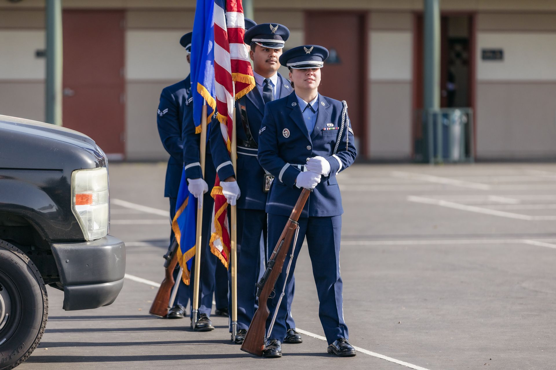 Uniformed honor guard standing with rifles and a U.S. flag in a parking lot