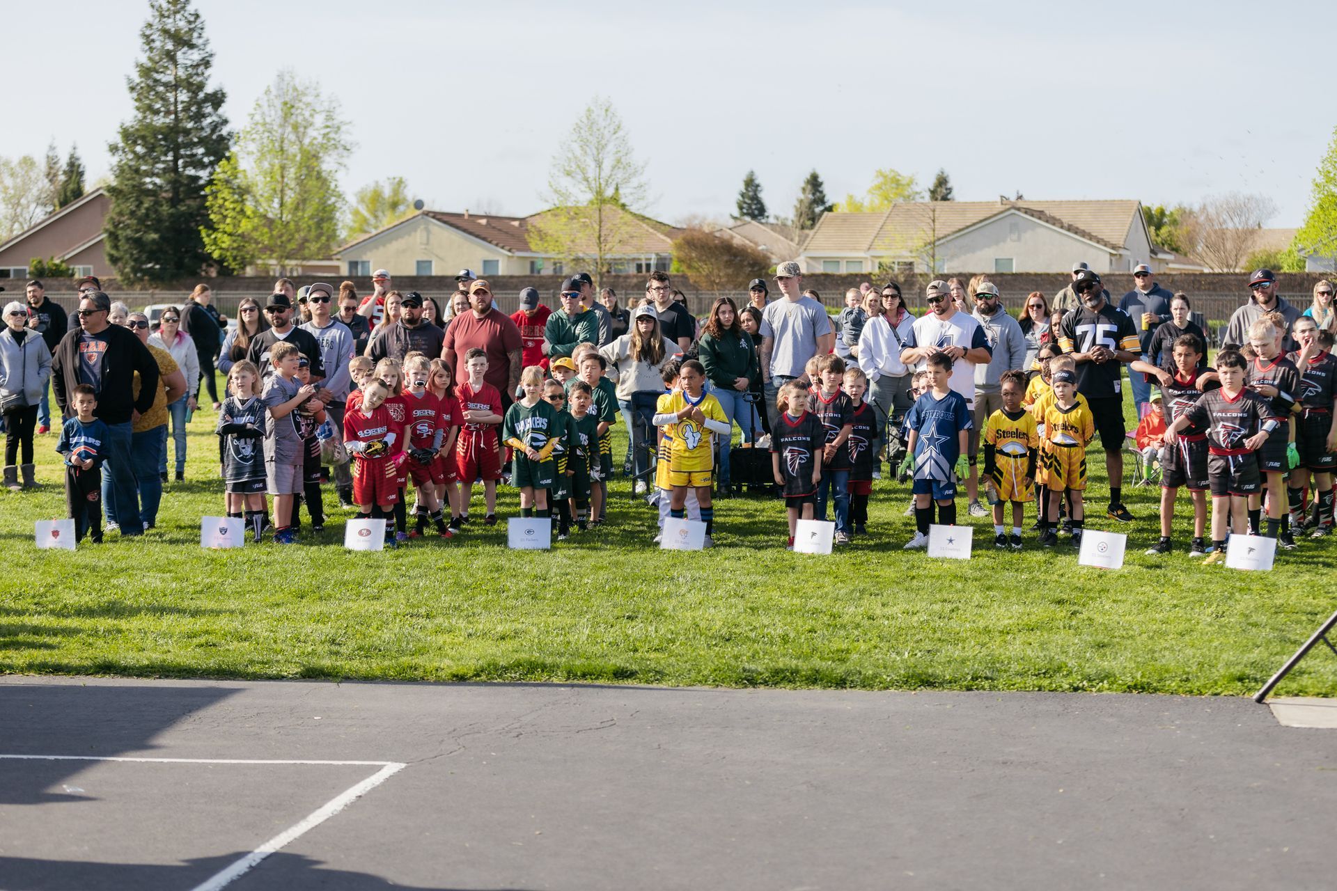 Large group of people standing on a grassy field in front of houses, holding signs at an outdoor event