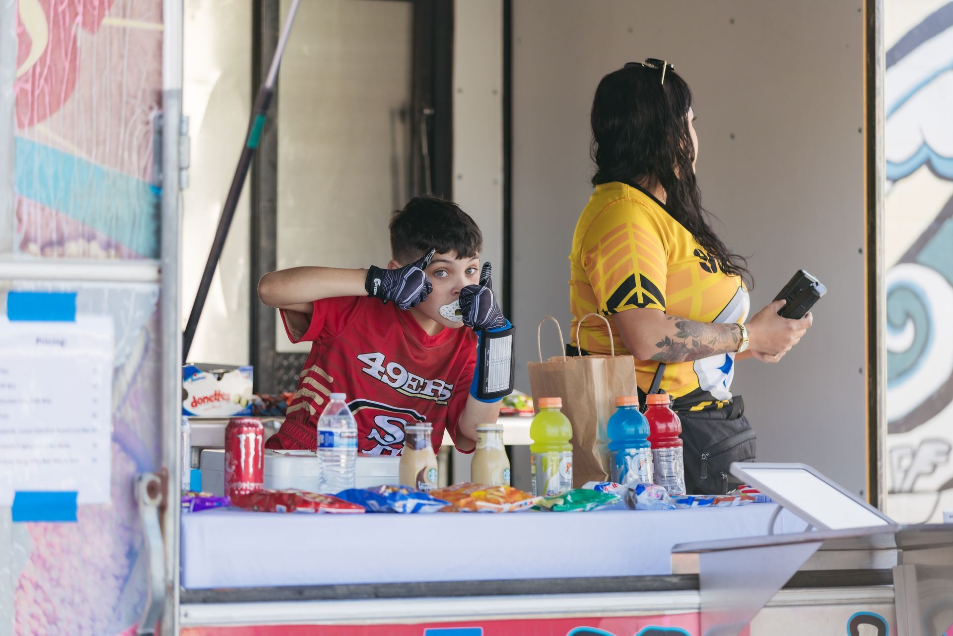 People at a table with drinks and snacks, one in a red shirt talking on the phone and another in yellow standing nearby.