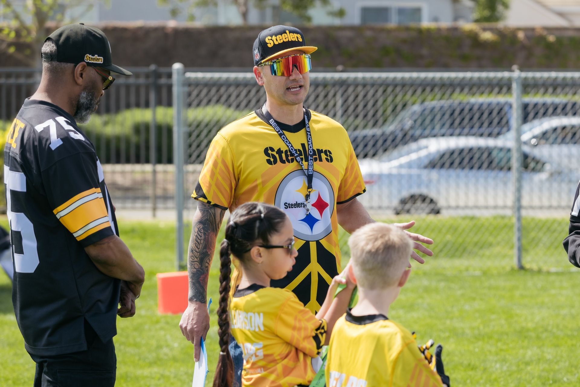People in yellow sports jerseys standing on a grassy field near a fence and parked cars