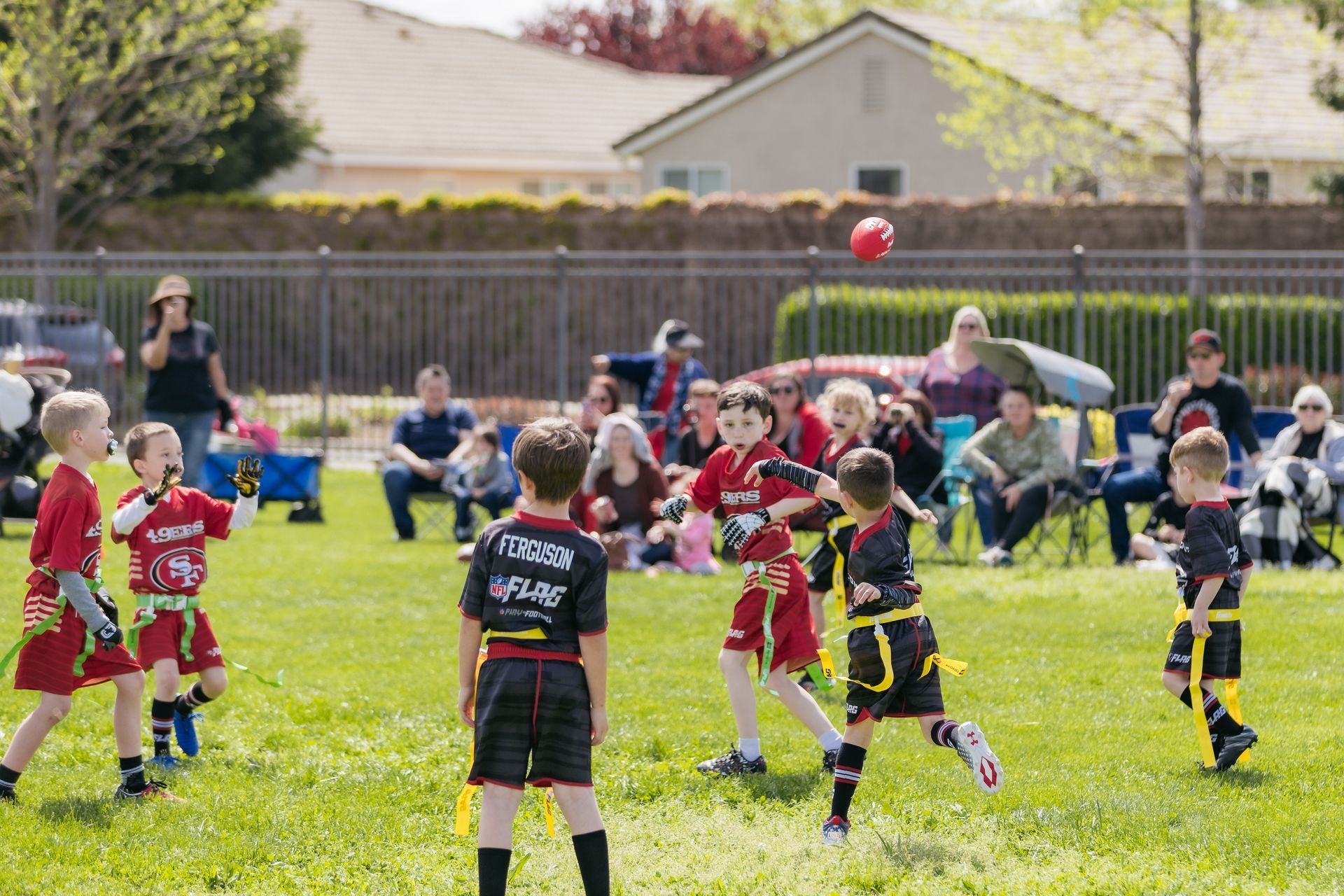 Youth soccer game on a grassy field, with children in red and black jerseys chasing a red ball.