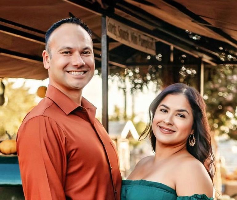 Two people smiling outdoors under a wooden roof, with warm sunset light in the background