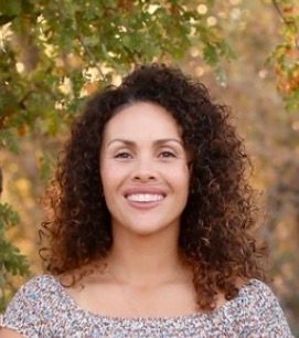 Smiling person with curly hair outdoors, standing before soft autumn foliage.