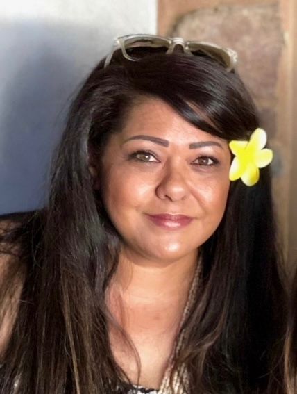 Woman with long dark hair, yellow flower in hair, and sunglasses on head, smiling indoors.