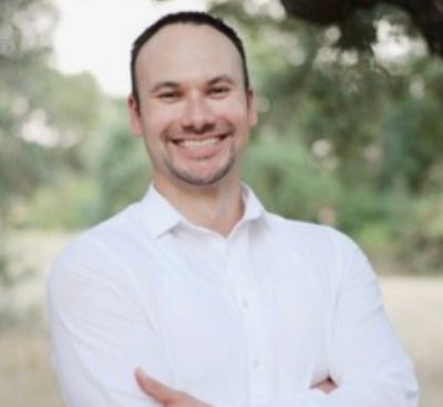 Smiling person in a white shirt standing outdoors with arms crossed.