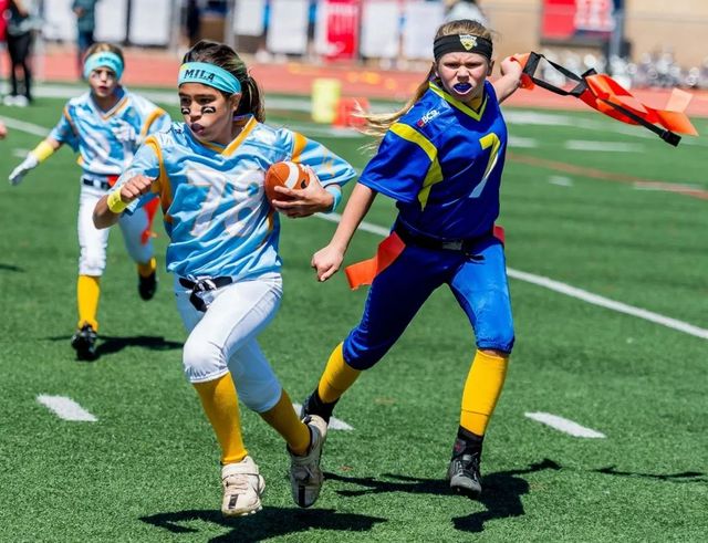 Youth flag football players sprint on a turf field, one carrying the ball while defenders chase.