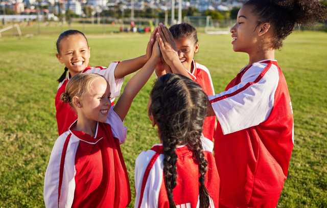 Children in red sports jerseys high-five on a grassy field during a game practice