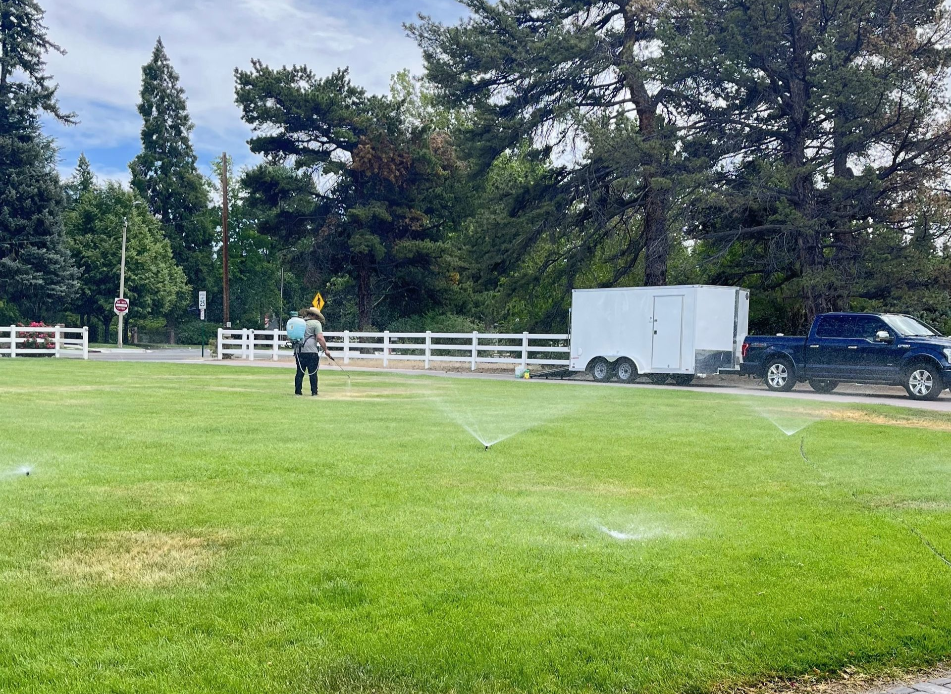 Person spraying a lawn, with sprinklers, trailer, and truck parked nearby.