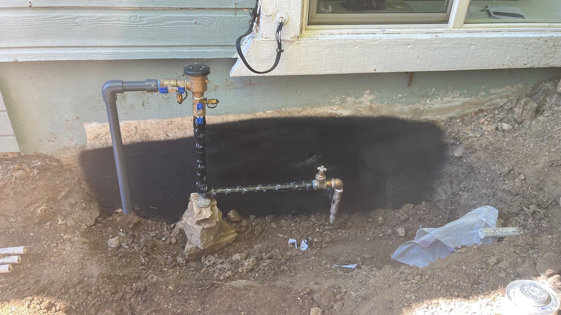 Outdoor water pipe and valve assembly with water spraying, against a wall and dirt.