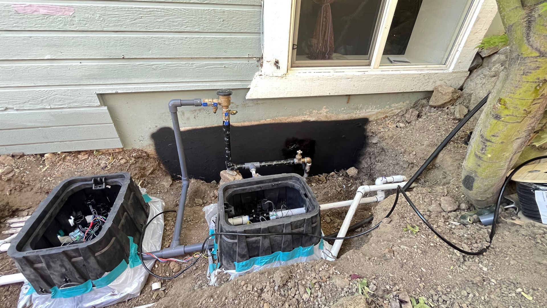 Irrigation system with two black valve boxes, pipes, and a water spigot near a building and tree.