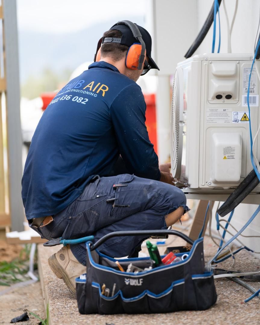 A Man Is Kneeling Down Next to A Tool Bag While Working on An Air Conditioner — Staub Air in Garbutt, QLD