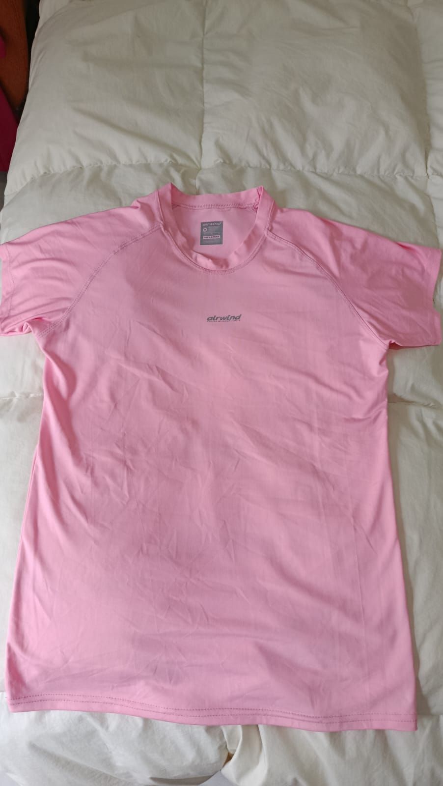 Camiseta rosa de manga corta extendida sobre una colcha blanca, con un pequeño logotipo en el pecho