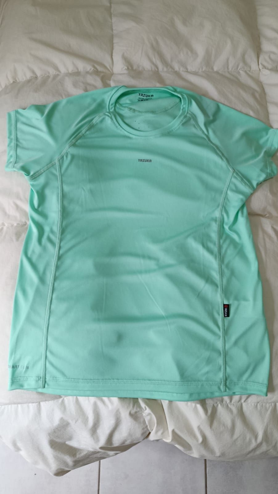 Camiseta rosa de manga corta extendida sobre una colcha blanca, con un pequeño logotipo en el pecho