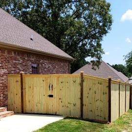 Wooden privacy fence and gate beside a brick house, with trees and blue sky overhead.