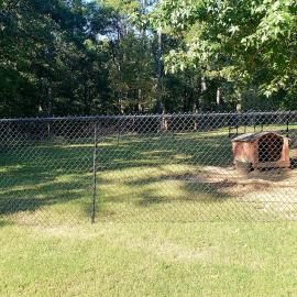 Chain-link fence around a grassy yard with a rust-colored doghouse and trees in the background