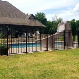 Outdoor swimming pool with a tan slide beside a building, viewed through a black fence.