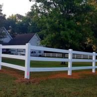 White ranch fence in a grassy yard with a house and trees in the background