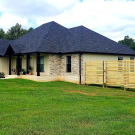 Single-story house with a dark roof, tan brick walls, and a fenced yard under cloudy skies