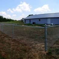 Chain-link fence around a grassy yard beside a long gray building under a partly cloudy sky