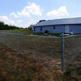 Chain-link fence in a dry yard beside a gray building under a blue sky.