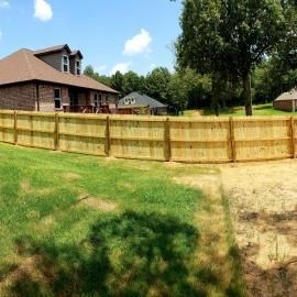 Backyard with wooden fence, green lawn, brick house, and large trees under a blue sky