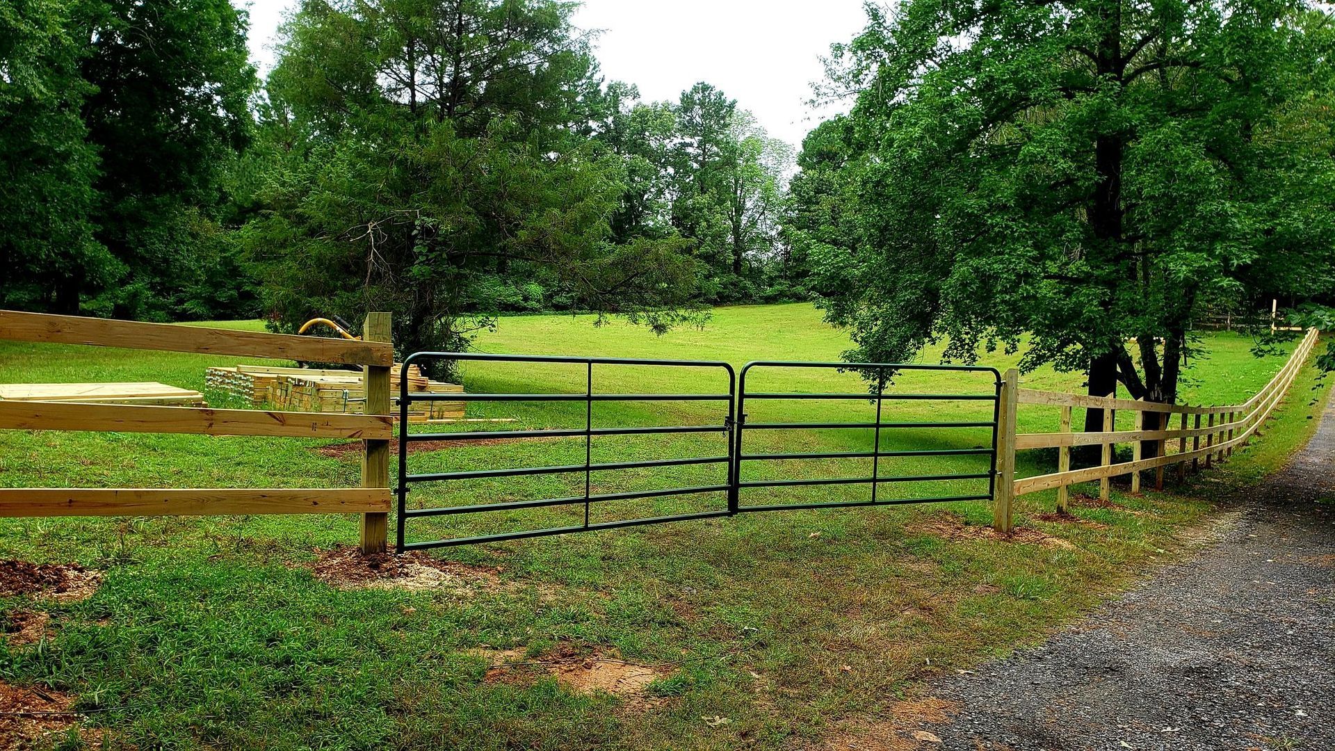 Black metal farm gate between wooden fences beside a gravel driveway, opening to a green field and trees.