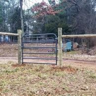 Metal farm gate between wooden posts on a grassy dirt path in a wooded area.