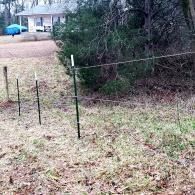 Wire fence posts along a grassy yard beside trees and a house in the background