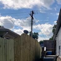 Narrow backyard alley with wooden fence, utility pole and wires under a partly cloudy sky