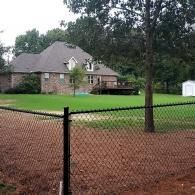 Large brick house behind a black fence, with a tree and lawn in a wooded suburban yard.