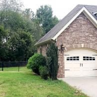 Suburban brick house with white garage door, green lawn, and trees by a fenced yard