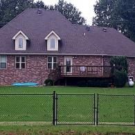 Brick house with dark roof, front deck, and fenced green lawn under cloudy sky