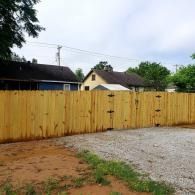 Yellow wooden fence with gate in a yard, with houses and trees in the background.