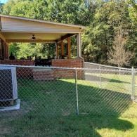 Chain-link fenced backyard with green grass and a covered patio beside trees