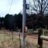 Rural roadside utility pole with an electrical box beside a dirt path and wire fence